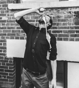 Black and white photo of a man spilling a drink from a can against a brick wall.