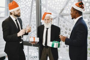 Three businessmen in Santa hats exchanging gifts and toasting at a holiday party indoors.