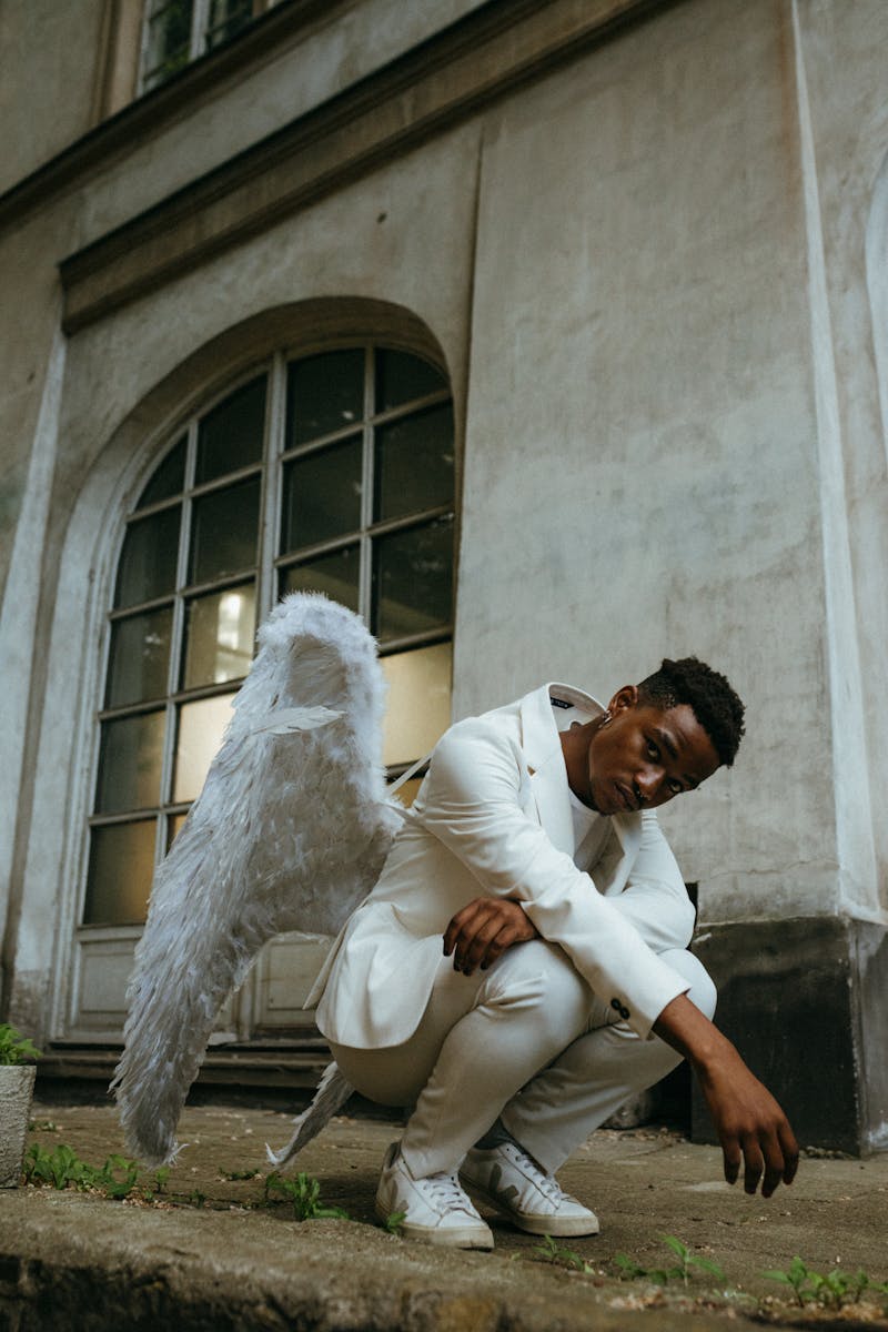 A man wearing a white suit with angel wings, sitting by an old building outdoors.