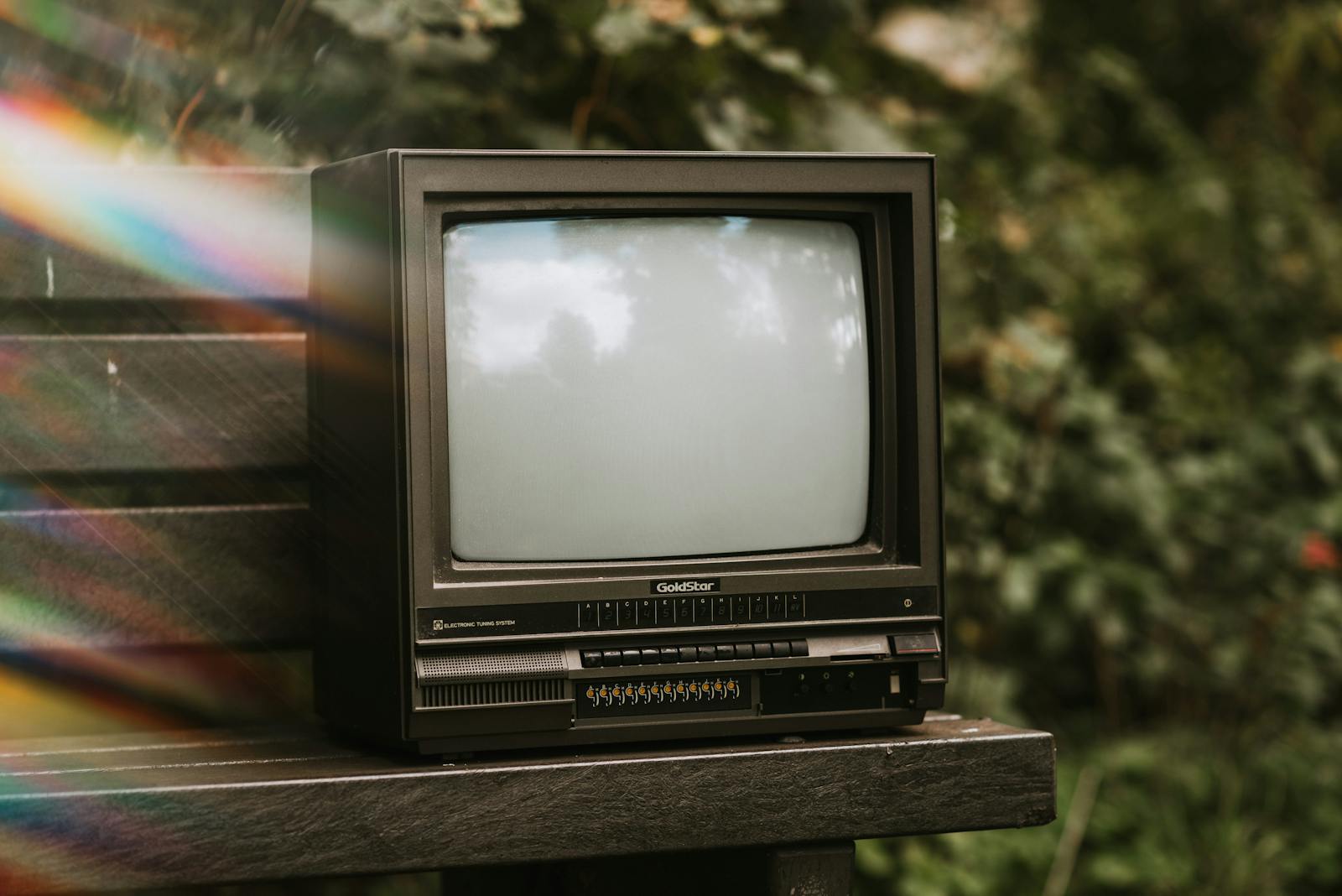 Abandoned vintage TV on a park bench surrounded by greenery, with artistic lens flare effect.