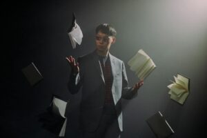 A man in eyeglasses and suit surrounded by floating books in a dramatic, dimly lit setting.