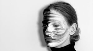 Black and white of contemplative young female model with painted face in turtleneck standing against light background